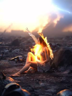 A surreal image depicts a woman with fiery wings and a glowing body, seated on a rocky, desolate terrain. The background suggests a dramatic sunset or sunrise, with swirling clouds and a bright light source. The overall composition evokes a sense of fantasy and struggle.