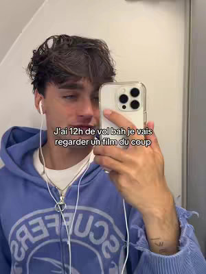 A young man with dark hair is taking a selfie holding a white phone with black camera housings. He is wearing a blue hoodie and has wired earbuds in. The background appears to be a plane interior.