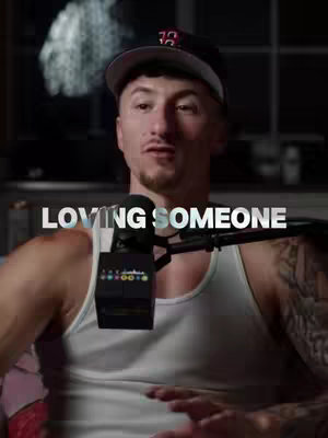 Image features a man in a baseball cap and a white tank top, centered and facing the camera. The background is out of focus. Overlayed text reads 'LOVING SOMEONE'.