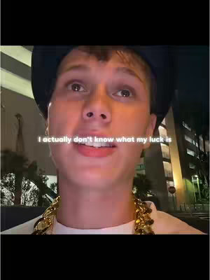 A medium shot features a young man in an urban setting. He has short hair and is wearing a black hat and a large gold chain. Text overlayed on the image reads "I actually don't know what my luck is". The background is blurred, suggesting a nighttime scene with buildings and streetlights.