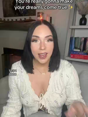 A young woman with dark hair and fair skin is centered in the frame, wearing a white, low-cut, knitted sweater. She is smiling and appears to be speaking. The background includes a fireplace, a ladder shelf with books, and neutral-toned walls. The overall composition is bright and inviting.
