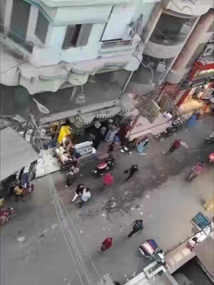 An aerial shot of a crowded city street. Buildings line the sides, with a few shops visible, and the street is filled with pedestrians, people on motorcycles, and some cars. Overcast lighting gives a muted color palette.