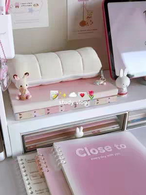 A neatly organized desk with various stationery items. In the foreground, a pink notebook, and some drawings with the text "Close to every day with you" on it. There is a small figure of a small bunny near a book. on the desk are posters and two miniature figures. The color palette is soft, dominated by pink and white tones, creating a cozy and inviting aesthetic.