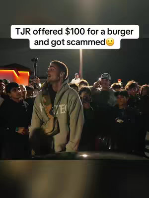 A nighttime shot of a crowd gathered around a man, the main subject, who is wearing a gray hoodie and facing away from the camera with a concerned expression. The image is brightly lit, with the subject and some of the crowd receiving direct light which gives clear silhouettes. A banner-like text overlay at the top of the frame gives context to the scene.