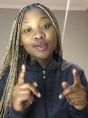A selfie-style shot features a young woman with braided blonde hair, looking directly at the camera with a slightly open mouth. She is pointing her index fingers towards the camera with both hands, and wears a black zip-up jacket. The background is a plain wall with a vertical light streak on the right.