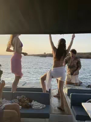 A group of people are on a boat on the water. Two women are dancing with open arms, with one leg raised, while another woman and an older man looks on. The sun is setting in the background.