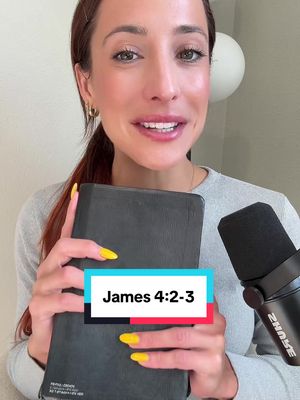 A woman with auburn hair holds a Bible open, looking at the camera and smiling. She has bright yellow nail polish and is wearing a gray long-sleeved shirt. In the background, there's a microphone and a white sphere. Overlaid on the image is a text box that says 'James 4:2-3'.