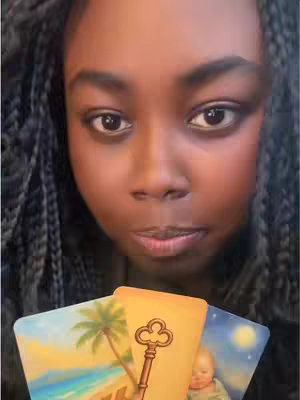 A close-up shot features a Black woman with her dark braids framing her face. Her eyes are directly facing the camera with a slightly tilted expression. She seems to be concentrating as she delicately holds three tarot cards in front of the camera. The cards themselves display a combination of images with light colour schemes. The setting is an indoor location, providing a simple, calm backdrop.