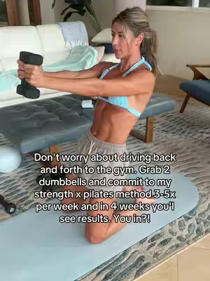 A woman in athletic wear is kneeling on a mat in what appears to be a home gym setting. She is holding a dumbbell in each hand and is framed in a medium shot. The background includes a sofa, a bench, and a rug.
