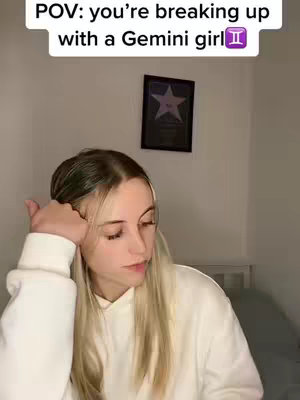A woman with long blonde hair is seated, resting her head on her left hand. She is wearing a white hoodie, facing slightly to the right. The background is a plain, light-colored wall with a framed picture hanging on it. The text overlay reads "POV: you're breaking up with a Gemini girl".