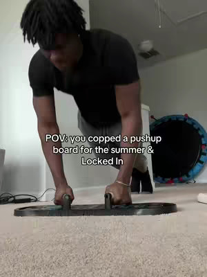 A person with dark hair doing a push-up on a black and gray push-up board, as if in home. The person is wearing a black shirt and light colored shorts.