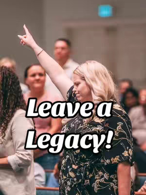 A woman in a black floral top raises her arm and points her index finger up in a large, brightly lit room. White text reading, "Leave a Legacy!" is overlaid on the image. Other people are visible in the background, out of focus.