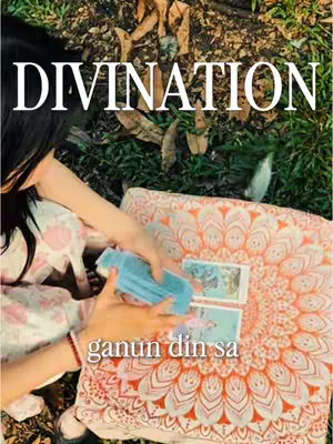 A woman is seated outdoors, hands arranging tarot cards on an intricately patterned, orange and white fabric. The word "DIVINATION" is in large white letters above her, set against a backdrop of grass, leaves, and a hint of sky. The overall composition is focused, and the color palette leans towards warm tones. The text "ganun din sa" is overlaid.