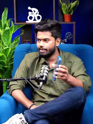 A man in a green shirt and jeans sits on a blue chair, talking while holding a blue phone. A microphone is positioned in front of him. The background includes a decorative white object and potted plants.