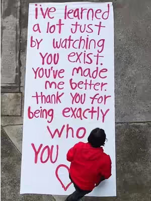 An overhead shot shows a person wearing a red hoodie and dark pants writing on a large white poster propped up on a gray surface. The poster contains handwritten text in red marker expressing gratitude. The person is seen from the back, their head and upper back visible as they write on the poster. The ground has grey pavers. A red heart is drawn at the bottom next to the word 'YOU'.