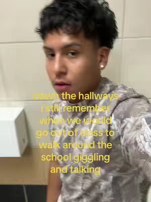 A young man is centered in a selfie-style video. He has short, dark, styled hair and a slightly concerned expression. He appears to be in a public restroom with off-white, tiled walls visible in the background. He is wearing a patterned, camouflage-style shirt and has a small stud earring in his right ear. Text overlays in yellow are over the image.