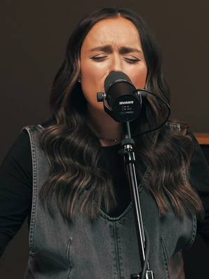 A woman with long brown hair, wearing a denim vest and a black top, sings into a microphone with her eyes closed, conveying deep emotion. The background is a muted, neutral color.