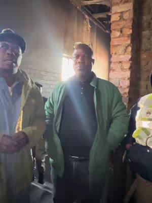 The image shows a scene inside a building, possibly dilapidated. Three men are visible, the closest two centered in the frame. The man in the middle, wearing a green jacket over a black shirt, is the main subject. The lighting is somewhat dim, with a bright light source behind the man, creating a glare. To the left, a man wears a hat and a jacket, while another man is shown to the right, slightly cut off. The interior reveals brick and building materials, and the overall composition emphasizes the environment.