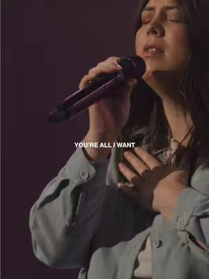 A woman with dark hair, wearing a gray top, is singing into a microphone, with her eyes closed and head tilted back. The background is dark and out of focus. Text overlay in white that says "YOU'RE ALL I WANT".