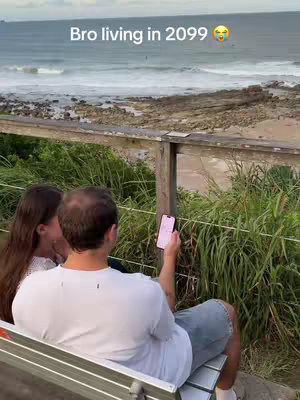A man and a woman sit on a bench overlooking a scenic view of the ocean. The man is holding a smartphone, and a text overlay indicates the video's theme.