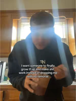 A person wearing a black hoodie is shown. The person is looking down with a slight glow on their head. The background consists of cabinets, a window, and blinds.