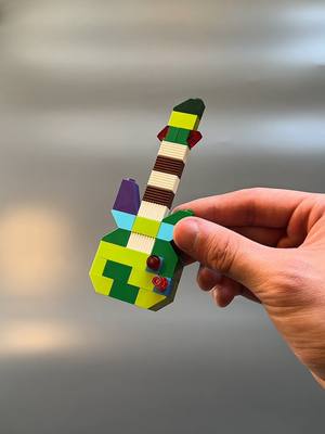 A hand holds a colorful guitar-shaped Lego model against a blurry gray background. The Lego guitar is the focal point, showcasing a design made from numerous colored bricks. The composition is simple, with the hand holding the model at an angle.