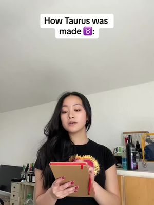 A young woman with long, dark hair is holding a notepad and looking at it attentively. The background is a nondescript room setting with white walls and minimal furnishings. Text overlaid on the top of the image reads: How Taurus was made ♉️.