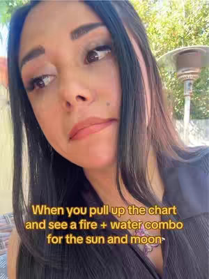 A woman with long black hair looks slightly to the left with a neutral expression. The image is taken from a closer perspective in a mostly outdoor setting. She is wearing a dark top, and text is overlaid at the bottom, relating to astrology and star signs.