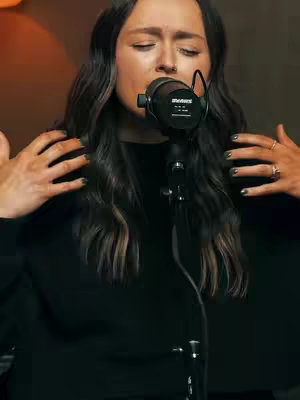 A woman with long, dark hair stands in front of a microphone, eyes closed, seemingly singing. Her arms are raised to the sides, hands visible, wearing rings. She's wearing a black garment.