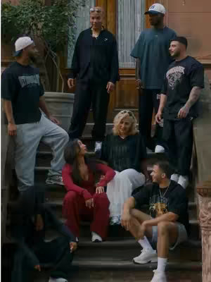 The image shows a group of people, mostly standing, on a set of outdoor stairs. A woman in the center, wearing a red outfit, is seated. The people are diverse in appearance and attire with a mix of casual and athletic wear.