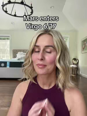The image shows a woman with blonde, wavy hair in a home setting. The text overlay says, 'Mars enters Virgo 6/17'. She is wearing a sleeveless burgundy top and has her eyes closed with a gentle expression, hands clasped in front. Behind her is a bedroom visible with a bed and art piece, typical of a warm, well-lit home.
