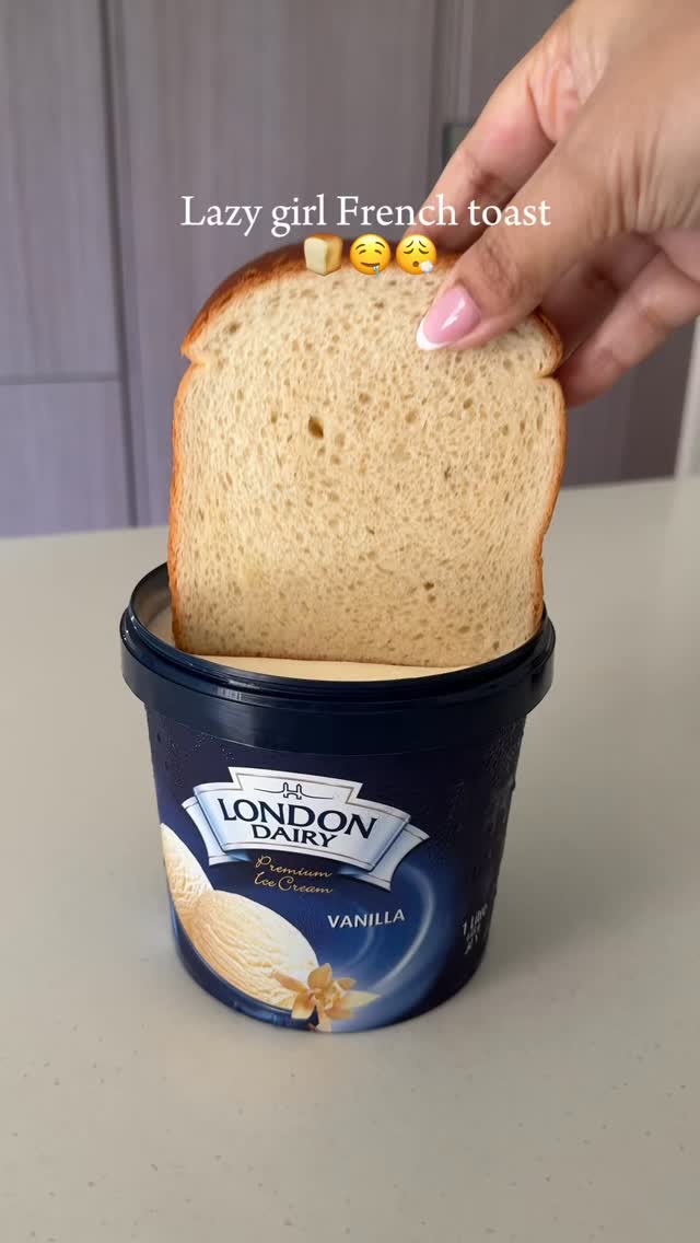 A hand holding a slice of bread dipped into a tub of vanilla ice cream. The bread is in the center, with the ice cream container below with the text \"London Dairy Vanilla\". The background has a neutral color.