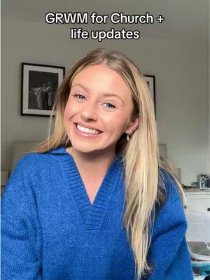 A woman with long blonde hair is smiling at the camera. She's wearing a blue sweater, and behind her are framed pictures on a white wall. The image is well-lit and appears to be taken indoors.