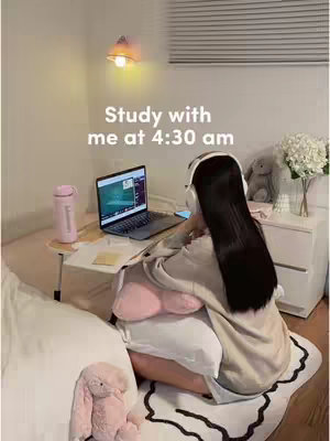 A woman with long black hair wearing white headphones is shown from the back, sitting on a bed and studying at a laptop. The room is softly lit, with a lamp, a water bottle, a stuffed animal and a flower vase on a nightstand. Text overlayed on the image reads, "Study with me at 4:30 am".