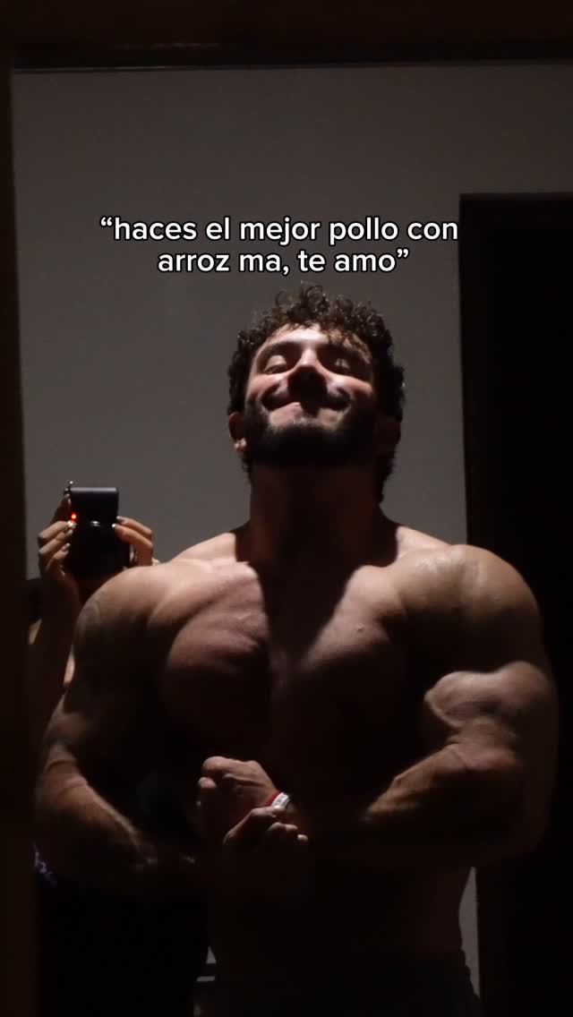 A muscular man is flexing his arms, showing his physique. A woman's hand holding the phone can be seen on the left to take a picture in a dark room. There is overlaid text at the top.