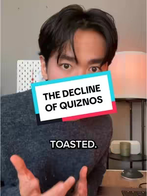 A person with dark hair wearing a gray sweater is the central focus, gazing at the camera. Overlaid on the image is a rectangular graphic with the words "THE DECLINE OF QUIZNOS" and "TOASTED" in white font, set against a background of teal, red, and white. The background includes a white board, a lamp, and a table with a keyboard.