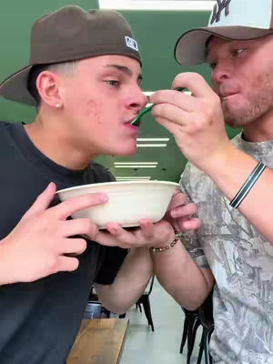 Two men are featured. One is wearing a brown cap and the other is wearing a light grey cap. The man in the brown cap is receiving a bite from the other man's spoon.