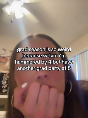 A selfie-style shot featuring a young woman from about the mid-chest up. She has long brown hair and is wearing hoop earrings. Her right hand is raised to her mouth, with her fingers partially covering her lips. The background is blurred, with a ceiling fan and a hint of a patterned surface visible. Text overlayed on the image reads, 'grad season is so weird because wdym i'm hammered by 4 but have another grad party at 8'.