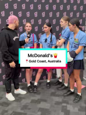 A man with pink hair is interviewing 5 teenagers outside. They're all smiling and seem interested in what the man is saying. A banner indicating where they are appears between their faces, reading "McDonald's Gold Coast, Australia". The background consists of a pattern of lowercase "g" characters.