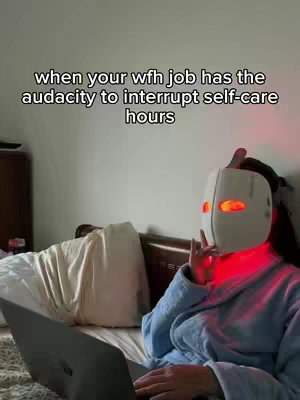 A woman is sitting in bed, seemingly in the middle of a work-from-home setup, with a laptop on her lap and a light therapy mask on her face. Overlayed text states the frustration of getting interrupted during self-care time.