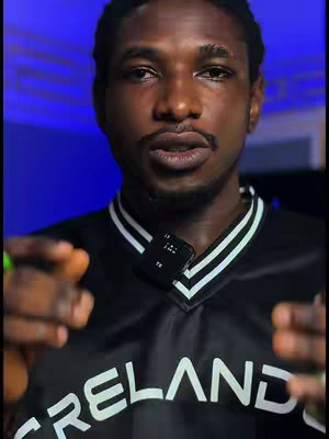 A man with a short beard and a serious expression is the focus of a medium shot. He is wearing a black jersey with white stripes on the collar and sleeves, with the word 'IRELAND' prominently displayed. A small microphone is clipped to his shirt. The background is a gradient of blue light.