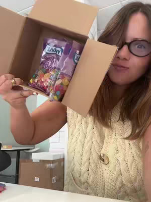 A woman with glasses holds open a cardboard box filled with bags of colorful candies, offering a view of the contents. She is wearing a cream crocheted vest and rings on her fingers, smiling at the camera. The background shows a partial view of an office-like setting.