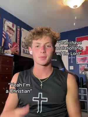 A young man with short, styled blonde hair wearing a black tank top with a cross pendant is centered in the frame. Text overlays include quotes and a cross symbol. The background features posters and a lamp.