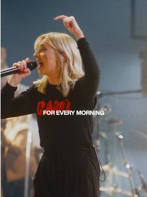 A woman with blonde hair is singing into a microphone, her right arm is raised. She wears black clothing with the words "GATHER" in red on the front. The background is out of focus with stage lighting present.