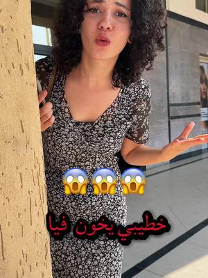 A woman with curly dark hair stands outdoors, her upper body visible. She looks directly at the camera with a slightly surprised or concerned expression, her lips pursed. She's wearing a black dress with a small, white floral pattern. Her right arm is bent, and her hand is gesturing to the side. A textured, tan surface, likely a stone pillar or wall, is to her left. Behind her is a darker background with a glimpse of a building and some greenery. The image contains text overlayed: Arabic text in the color red and 3 emoji faces with shocked expressions and there is a sign visible in the background.