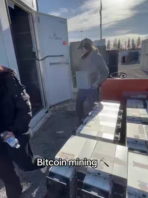 A person in a cap and jacket stands near an open white storage container, with several other similar containers and a glimpse of a blue sky in the background. Along the bottom of the image, the text "Bitcoin mining" is displayed.