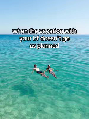 Aerial view of a serene ocean scene. A man and a woman are floating on their backs in the clear, turquoise water. The sky is a vibrant blue, providing a contrasting backdrop. The composition is symmetrical, with the subjects centered.