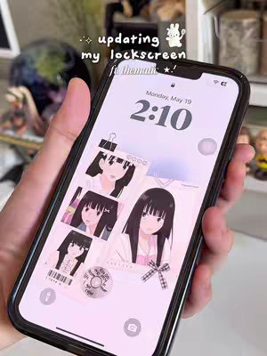 A hand holding a phone displaying a custom lock screen featuring various anime-style illustrations of a girl with long, black hair. The background is a light pink color. The lock screen also displays the current date and time. The phone is held in front of a blurred background that suggests a room with various objects.