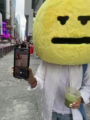 A person in a white top holds a phone up to film. The person is wearing a large yellow emoji-like head, possibly a costume. They are standing on a city sidewalk with a visible street and blurred buildings in the background. A green drink is in the person's hand.