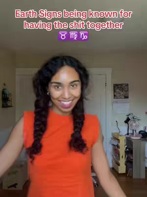 A woman smiles at the camera against a neutral wall, wearing a sleeveless orange dress and her dark hair in two braids. Text overlayed on the image declares: Earth Signs being known for having the $hit together. Zodiac symbols follow. The lighting is natural, and the setting appears to be a home or studio. The frame is a medium shot, capturing the woman from the chest up.